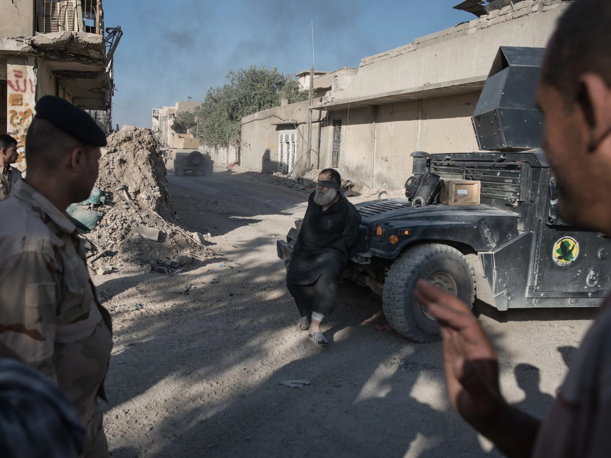 A blindfolded man with a white beard and his hands behind his back sitting against an armoured vehicle on a damaged street while soldiers watch
