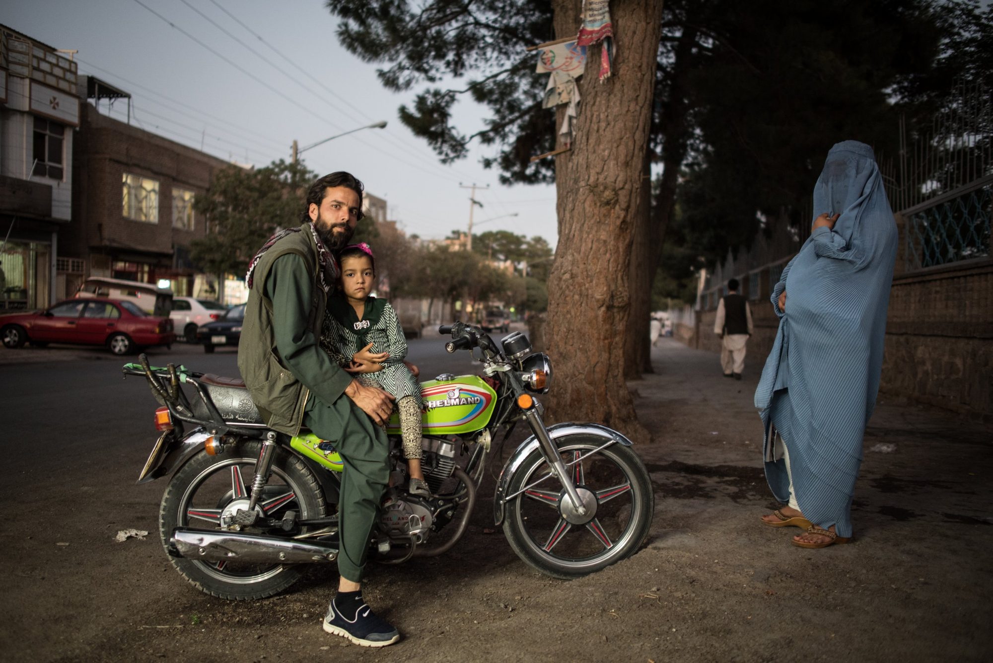 A man and a young girl sitting on a green motorcycle on a street, with a woman in a blue burqa standing nearby
