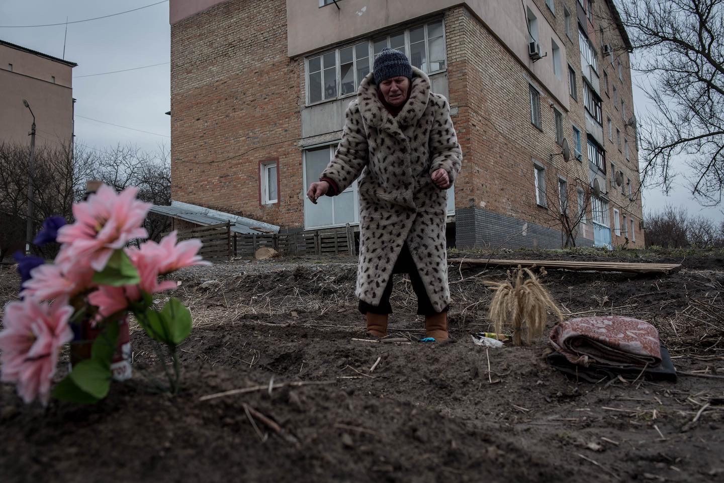 A woman in a leopard-print coat standing over a freshly dug grave in front of an apartment block