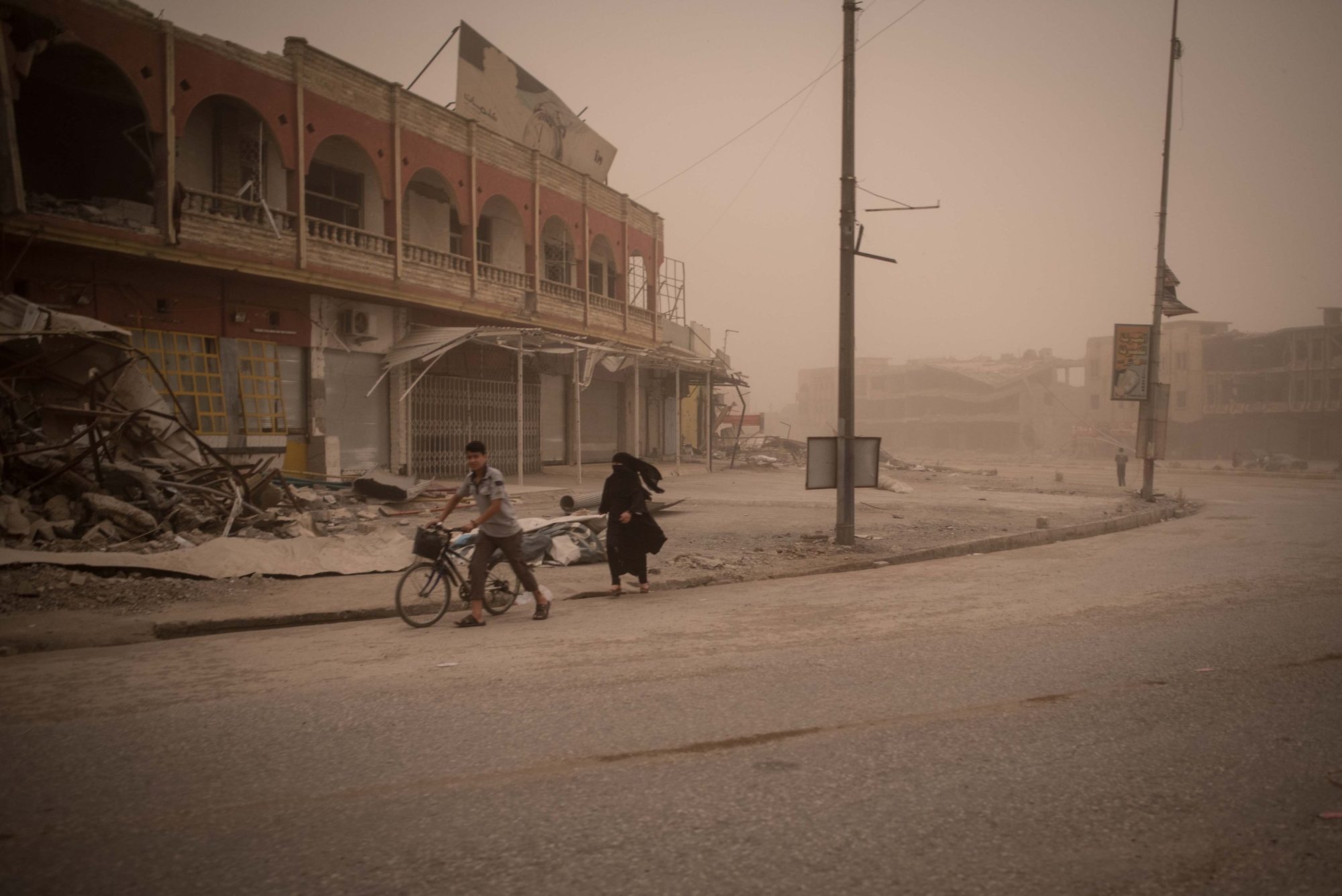 A boy pushing a bicycle and a woman in black walking through a dust storm past damaged buildings