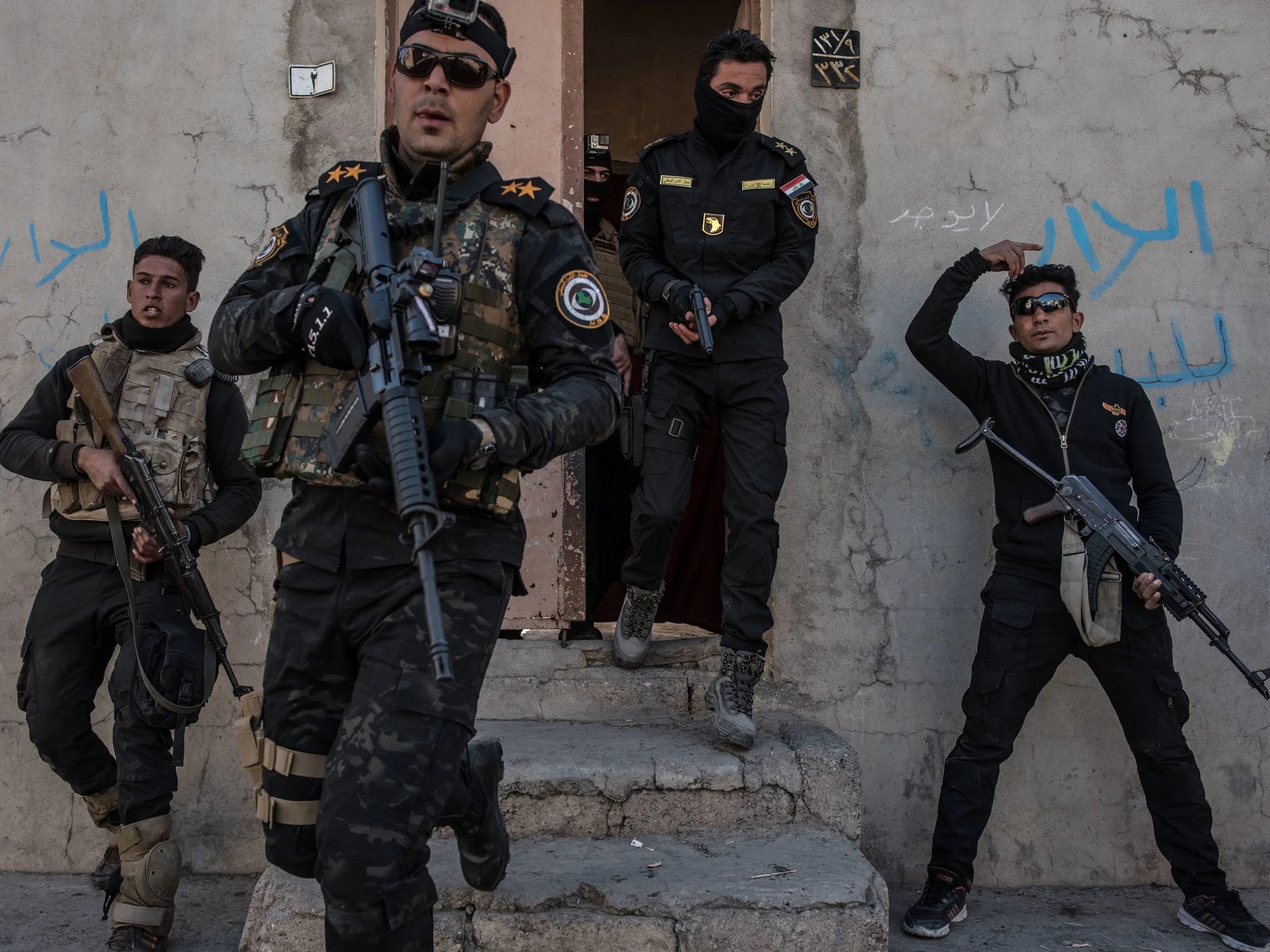 Armed men in black uniforms and tactical vests stepping out of a doorway and standing against a wall with Arabic graffiti